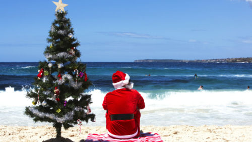 Man dressed as Santa Claus sitting by Christmas tree on beach, rear view